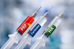 Three syringes labeled PRP, Cortisone, and HA, filled with red, blue, and clear liquids, arranged side by side against a blurred blue and white background, representing different joint pain injections
